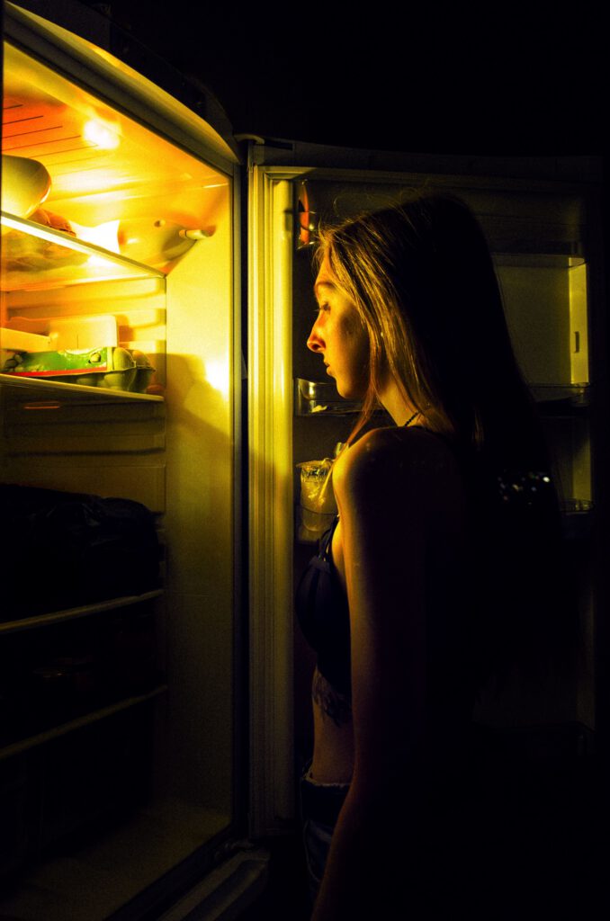 Woman standing at an open refrigerator at night representing automatic eating habits